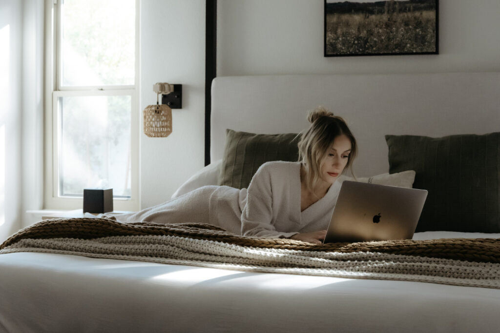 Luxury wedding photographer Kait working on her laptop while relaxing in a bright, modern hotel bedroom.