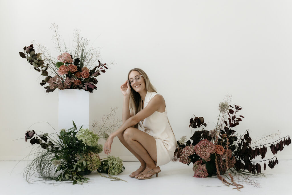 A smiling blonde female entrepreneur in a cream sleeveless blazer dress and clear glasses, crouching gracefully in a bright studio surrounded by organic floral arrangements of roses, hydrangeas, and dark fall foliage.