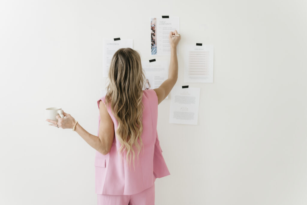 Rear view of a blonde female entrepreneur in a pink vest and matching pants holding a coffee mug while mapping out strategy, writing on printed brand pages taped to a minimalist white wall.