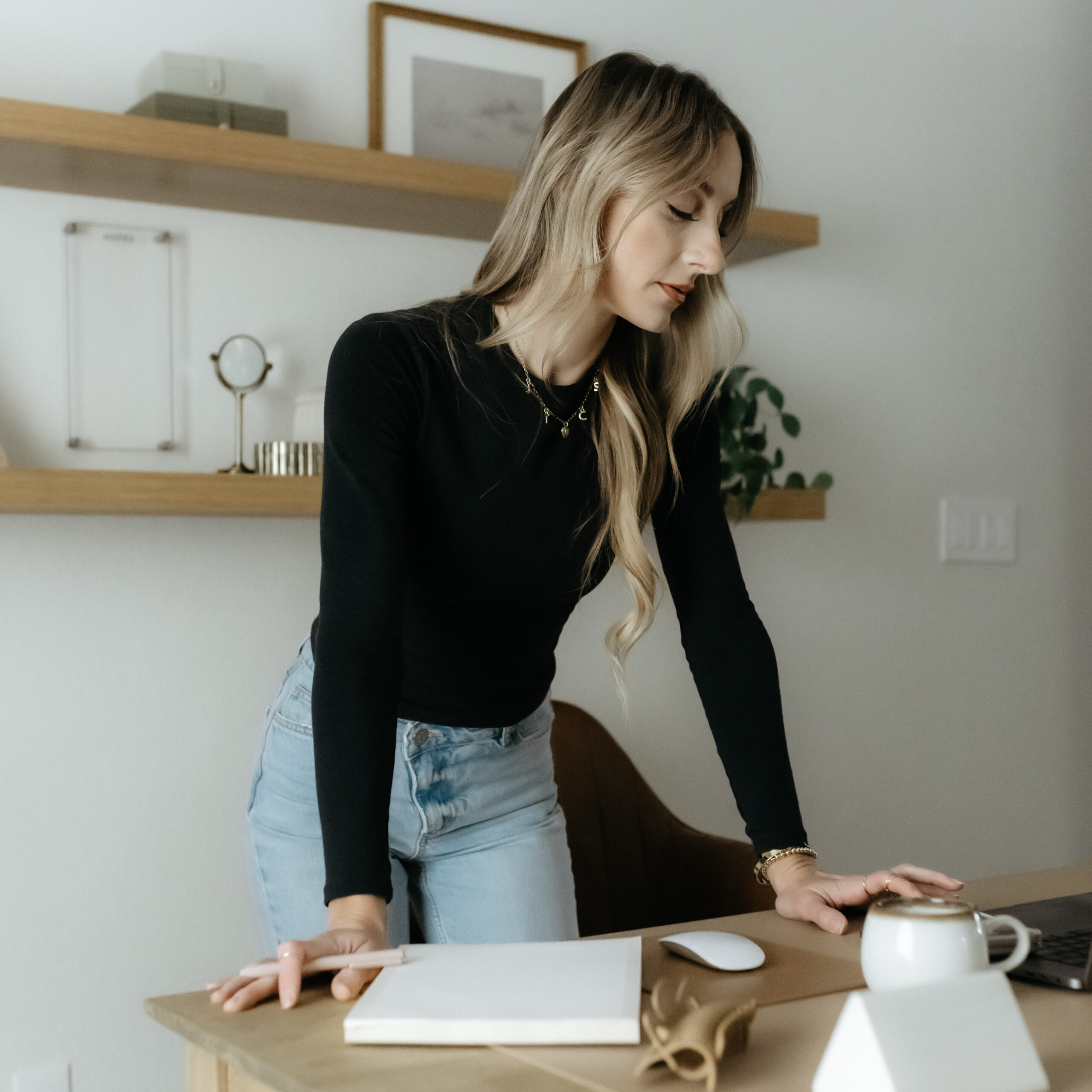 Creative business coach Kait working at a tidy wooden desk in a modern home office with floating shelves.